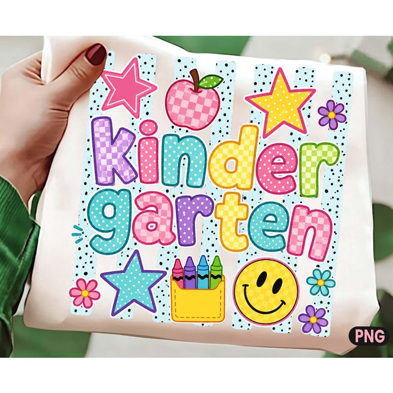 May include: A white t-shirt featuring a colorful graphic with the text Kindergarten in a playful font. The design includes a checkered apple, stars, flowers, a yellow pocket with crayons, and a yellow smiley face over light blue vertical stripes.