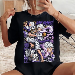 Puede incluir: Camiseta negra con un gráfico de estilo anime. El diseño presenta un personaje con ropa blanca en poses dinámicas, rodeado de detalles morados y amarillos. El texto "JOY BOY!!" es claramente visible.