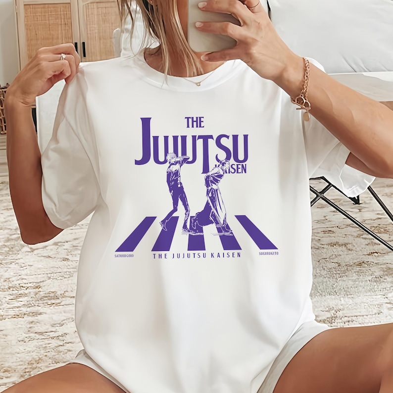 May include: White t-shirt featuring a purple graphic design. The design includes the words "THE JUJUTSU KAISEN" above an illustration of two figures on a crosswalk. The names "SATORUGOJO" and "SUGURIGETO" are printed below.