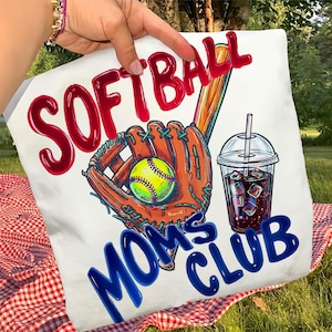Puede incluir: Cojín blanco con un diseño temático de softbol. El diseño incluye la palabra "SOFTBALL" en rojo, un guante de béisbol, una pelota de softbol, una bebida y las palabras "MOMS CLUB" en azul.