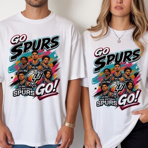May include: White t-shirts with a colorful graphic featuring the text "GO SPURS" and a team logo. The design includes images of basketball players and the words "SAN ANTONIO SPURS GO!"