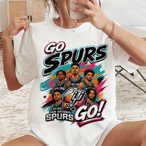 May include: White t-shirt featuring a graphic of the San Antonio Spurs basketball team. The design includes the words "GO SPURS GO!" in a bold font, along with images of the players and the team logo.