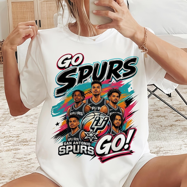 May include: White t-shirt featuring a graphic of the San Antonio Spurs basketball team. The design includes the words "GO SPURS GO!" in a bold font, along with images of the players and the team logo.