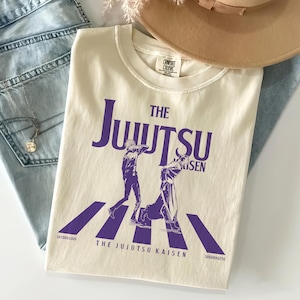 May include: Cream-colored t-shirt featuring a purple graphic of two figures on a crosswalk with the text "THE JUJUTSU KAISEN". The shirt is made of a soft material and is a comfortable fit. The shirt is paired with light blue jeans and a tan hat.