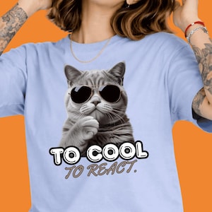 May include: Light blue t-shirt featuring a gray cat wearing sunglasses. The cat is depicted with a thoughtful expression. Below the cat is the text "TO COOL TO REACT."