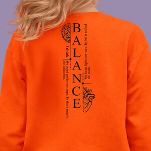 Peut inclure: Sweat-shirt orange vif avec le mot "BALANCE" en design vertical. Le motif comprend un cerveau, du texte et un cœur. Le texte se lit comme suit : "My heart lights my way. To feel is to find the truth."