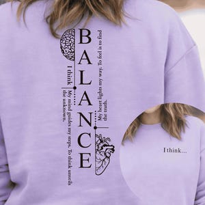 Peut inclure: Un sweat-shirt lilas avec le mot "BALANCE" imprimé verticalement en noir. Le design comprend des graphiques de cerveau et de cœur, avec le texte : "Je pense... Mon esprit guide mes pas. Penser révèle l'inconnu."