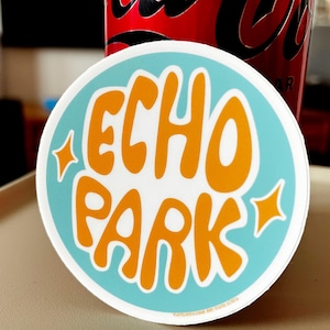 May include: A round sticker with the words "ECHO PARK" in orange, stylized lettering, set against a light blue background. The sticker has orange starburst accents and a white border. A red can is visible in the background.