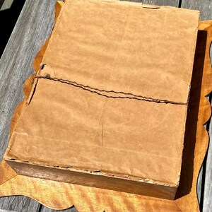 May include: A vintage framed artwork, viewed from the back. The frame is made of light-colored wood with a scalloped edge. The artwork is covered with a brown paper backing, secured with wire. The frame is rectangular and sits on a wooden surface.