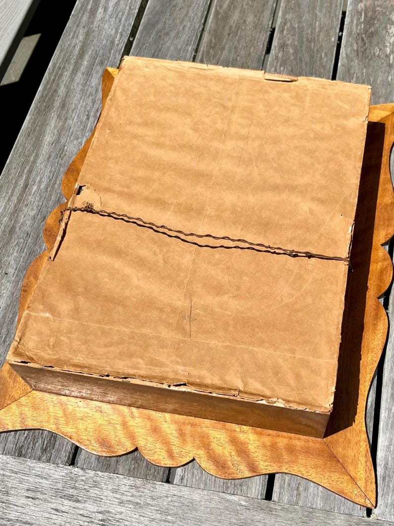 May include: A vintage framed artwork, viewed from the back. The frame is made of light-colored wood with a scalloped edge. The artwork is covered with a brown paper backing, secured with wire. The frame is rectangular and sits on a wooden surface.