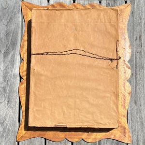 May include: Back of a framed artwork with a scalloped wooden frame. The frame is a light brown color. The artwork is covered with brown paper and secured with wire.