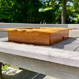 May include: A rectangular wooden box with a light brown finish. The box has a lid with a cutout design and is sitting on a weathered gray wooden surface. The background features lush green foliage.