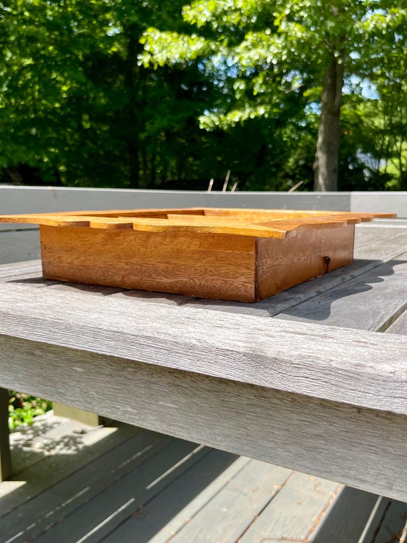May include: A rectangular wooden box with a light brown finish. The box has a lid with a cutout design and is sitting on a weathered gray wooden surface. The background features lush green foliage.