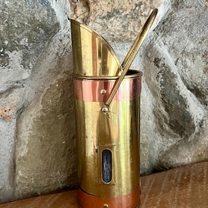 May include: A vintage brass coal scuttle with a copper band and handle. The scuttle has a cylindrical body with a small rectangular window and a scoop-shaped top. The handle is arched and attached to the top. The scuttle is displayed on a wooden surface.