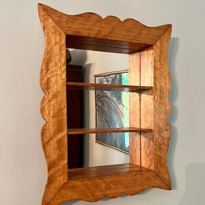 May include: A wooden wall-mounted shelf with a mirror backing. The shelf has three tiers and a decorative, scalloped frame. The wood has a warm, natural finish. The mirror reflects a painting and other objects.