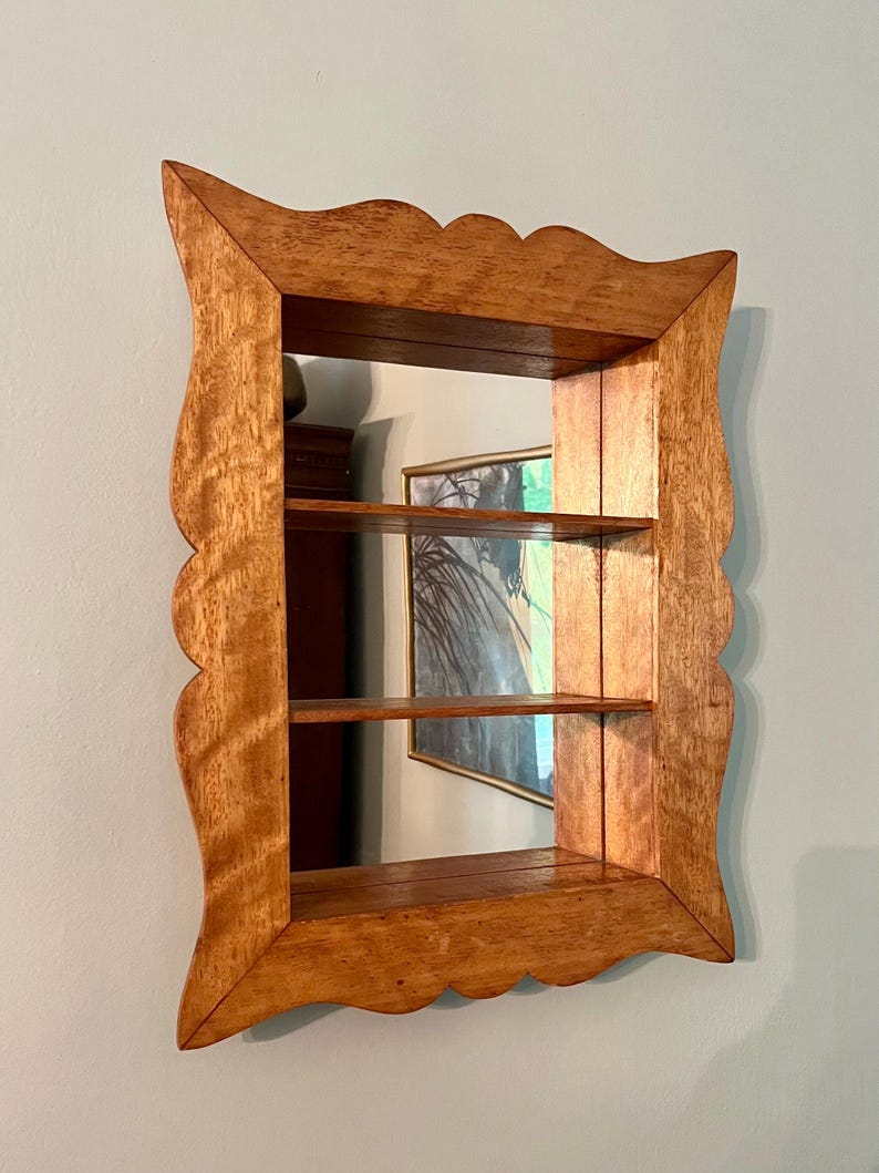 May include: A wooden wall-mounted shelf with a mirror backing. The shelf has three tiers and a decorative, scalloped frame. The wood has a warm, natural finish. The mirror reflects a painting and other objects.