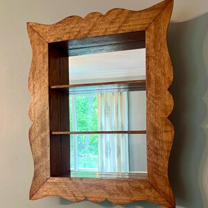May include: A wooden wall-mounted mirror with a scalloped frame. The mirror has two shelves and reflects a window with curtains. The wood has a warm, natural tone, and the frame has a decorative edge.
