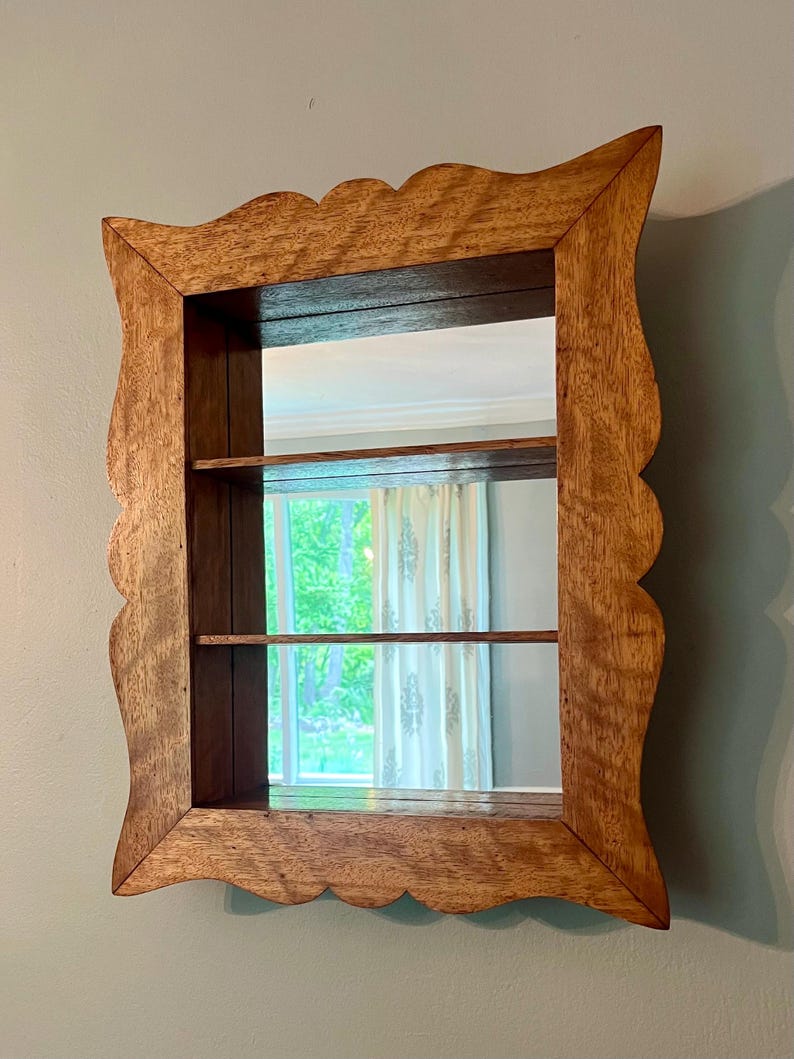 May include: A wooden wall-mounted mirror with a scalloped frame. The mirror has two shelves and reflects a window with curtains. The wood has a warm, natural tone, and the frame has a decorative edge.