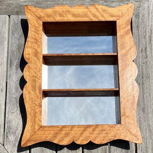 May include: A wooden shadow box with a scalloped edge and three interior shelves. The frame is a warm brown color, and the shelves are visible through the glass. The shadow box is designed to display small items.