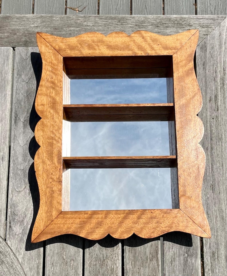 May include: A wooden shadow box with a scalloped edge and three interior shelves. The frame is a warm brown color, and the shelves are visible through the glass. The shadow box is designed to display small items.