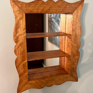 May include: A wooden wall shelf with a mirror backing and two shelves. The frame has a scalloped edge and is made of light brown wood. The shelves are also made of wood and are visible through the mirror.