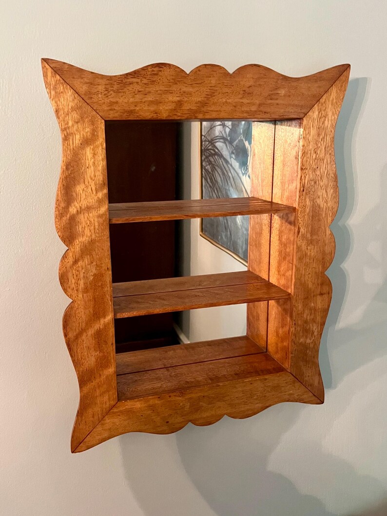 May include: A wooden wall shelf with a mirror backing and two shelves. The frame has a scalloped edge and is made of light brown wood. The shelves are also made of wood and are visible through the mirror.
