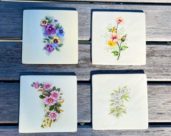 May include: Four square ceramic coasters with floral designs. Each coaster features a different arrangement of flowers in shades of purple, pink, yellow, and white. The coasters are set on a weathered wooden surface.