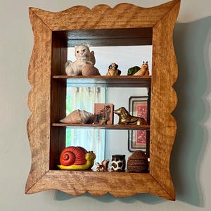 May include: A wooden shadow box with a scalloped edge, displaying various ceramic figurines on three shelves. The figurines include a cat, dogs, a snail, and other decorative items. The box is made of light brown wood and hangs on a light blue wall.