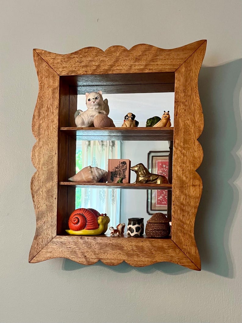 May include: A wooden shadow box with a scalloped edge, displaying various ceramic figurines on three shelves. The figurines include a cat, dogs, a snail, and other decorative items. The box is made of light brown wood and hangs on a light blue wall.