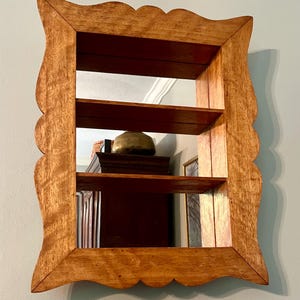 May include: A wooden wall-mounted mirror with two shelves. The frame has a scalloped edge and a natural wood finish. The mirror reflects the interior of a room, including a dark wooden cabinet and a brass-colored object.