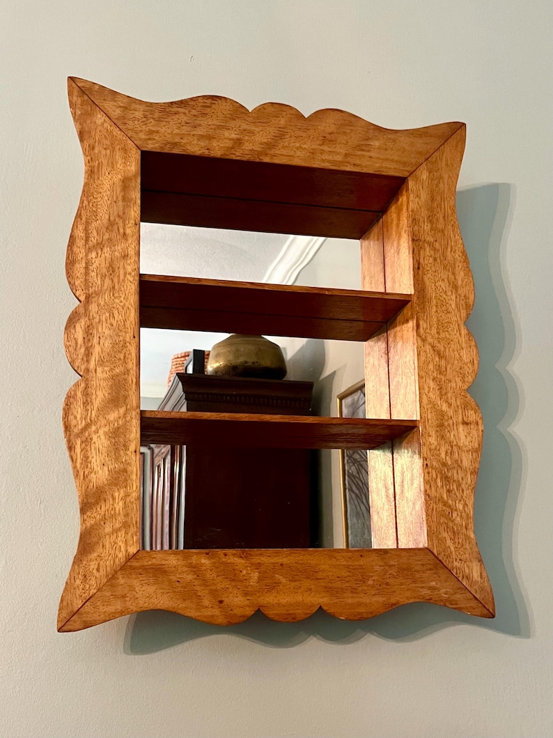 May include: A wooden wall-mounted mirror with two shelves. The frame has a scalloped edge and a natural wood finish. The mirror reflects the interior of a room, including a dark wooden cabinet and a brass-colored object.