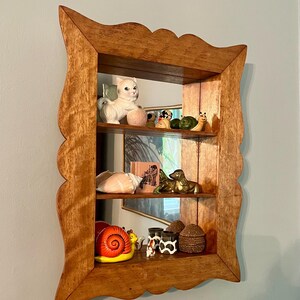 May include: A wooden shelf with a scalloped edge, displaying various figurines. The shelf has three levels, showcasing a white cat, a snail, a seashell, and other decorative items. The wood has a warm, natural tone.
