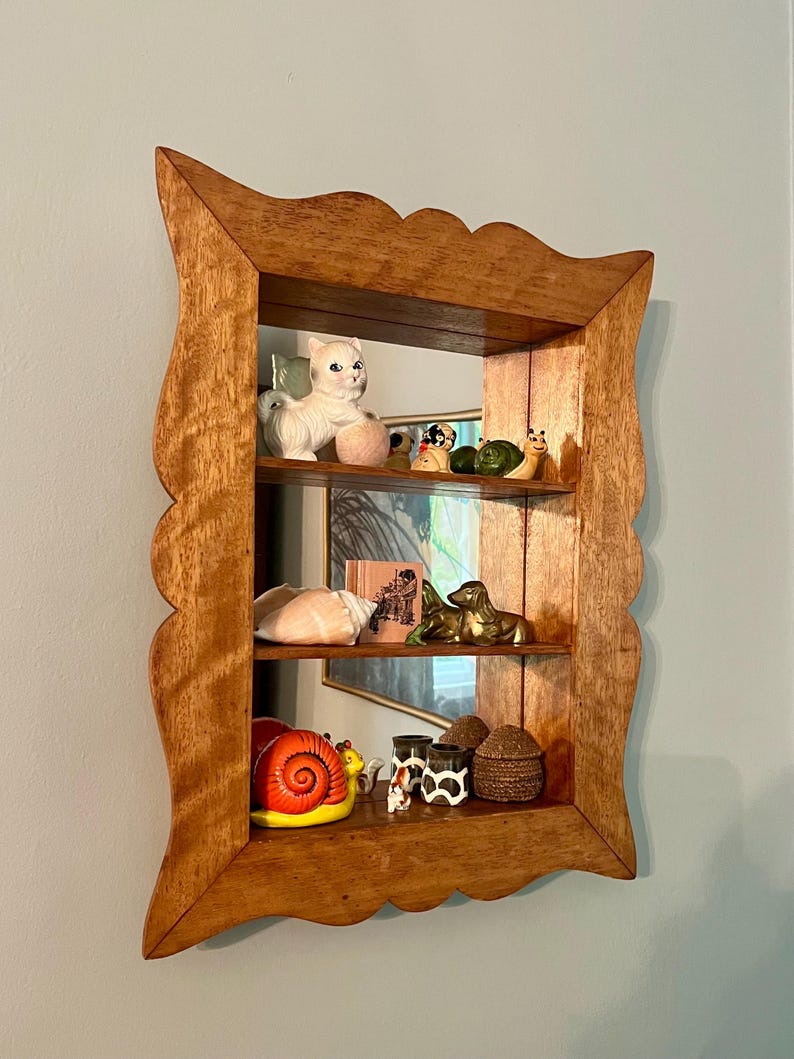 May include: A wooden shelf with a scalloped edge, displaying various figurines. The shelf has three levels, showcasing a white cat, a snail, a seashell, and other decorative items. The wood has a warm, natural tone.