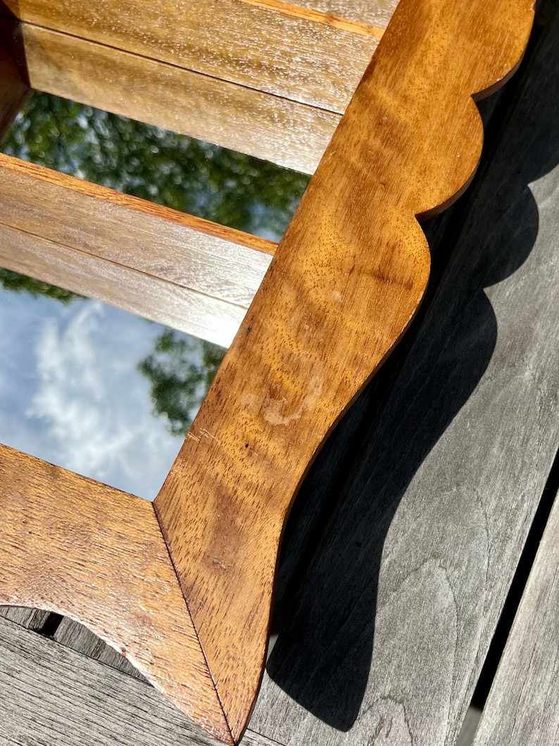 May include: A wooden framed mirror with a scalloped edge. The frame is a warm brown color with visible wood grain. The mirror reflects a view of the sky and trees. The mirror is rectangular.