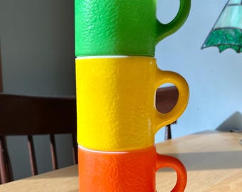 May include: Three brightly colored mugs stacked on top of each other. The top mug is green, the middle is yellow, and the bottom is orange. Each mug has a textured surface and a curved handle.