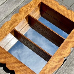 May include: A wooden wall mirror with a scalloped edge and a rectangular shape. The mirror has two wooden dividers and reflects the sky. The wood has a warm, natural tone.