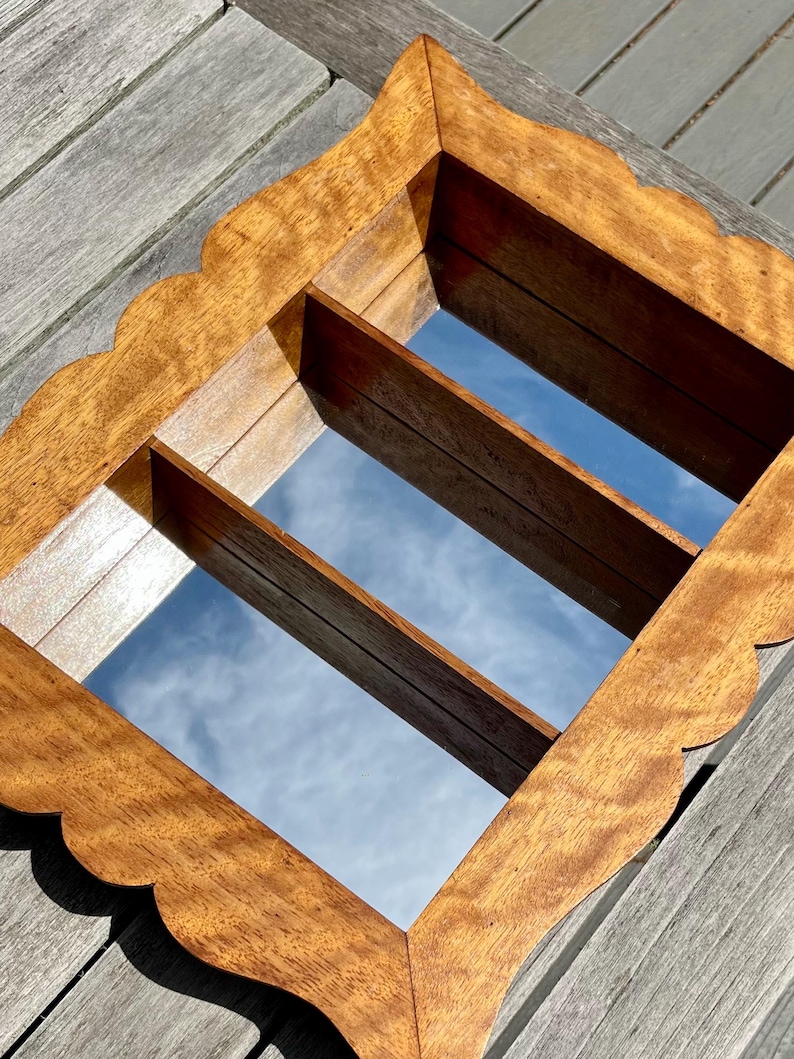 May include: A wooden wall mirror with a scalloped edge and a rectangular shape. The mirror has two wooden dividers and reflects the sky. The wood has a warm, natural tone.
