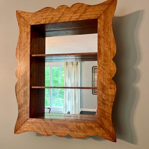 May include: Wooden wall-mounted mirror with a scalloped frame. The mirror has two shelves and a rectangular shape. The wood has a natural finish, and the mirror reflects a window and interior space.