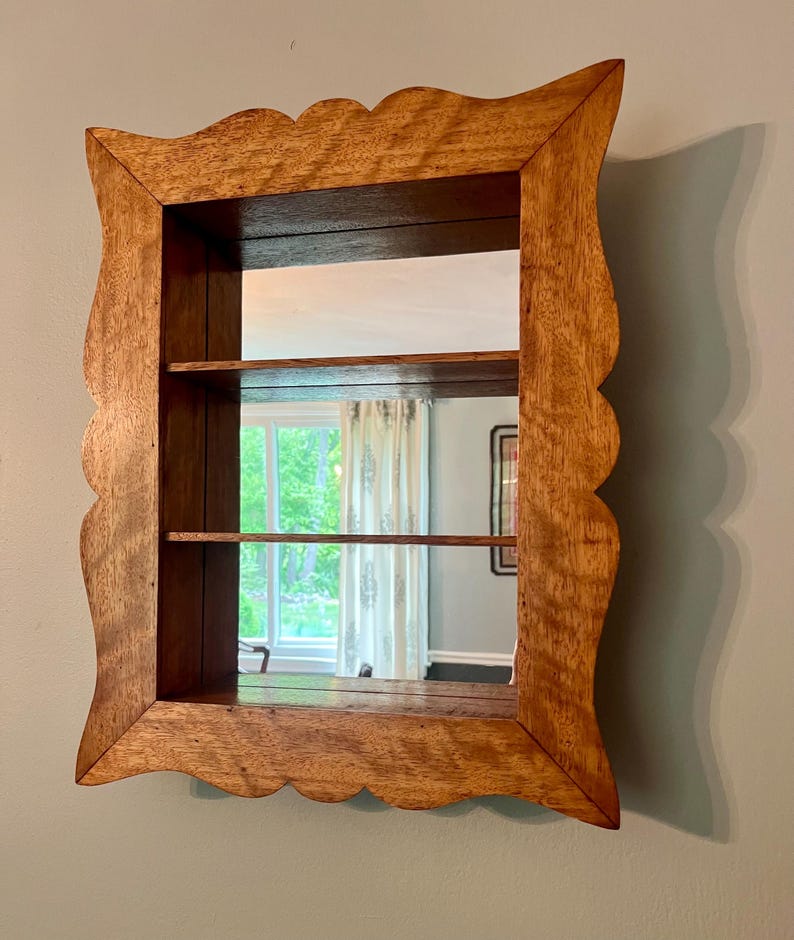 May include: Wooden wall-mounted mirror with a scalloped frame. The mirror has two shelves and a rectangular shape. The wood has a natural finish, and the mirror reflects a window and interior space.