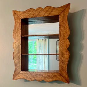 May include: Wooden framed mirror with a scalloped edge. The mirror has two shelves inside the frame. The wood has a natural finish. The mirror is mounted on a light blue wall.