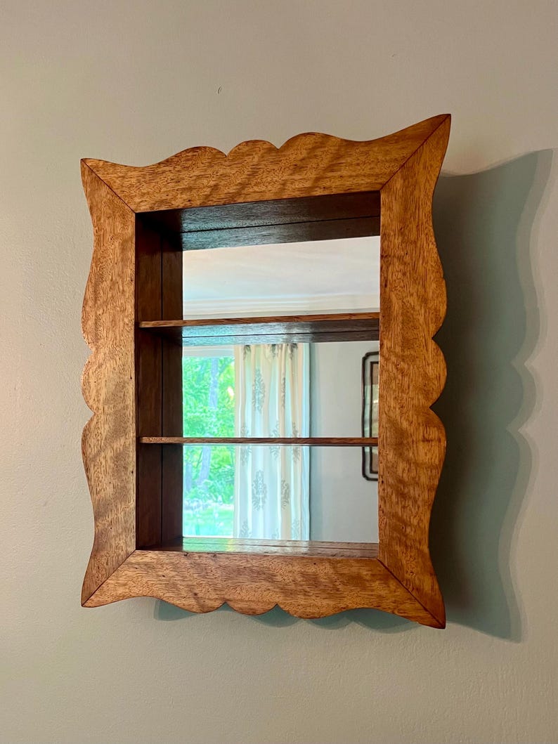 May include: Wooden framed mirror with a scalloped edge. The mirror has two shelves inside the frame. The wood has a natural finish. The mirror is mounted on a light blue wall.