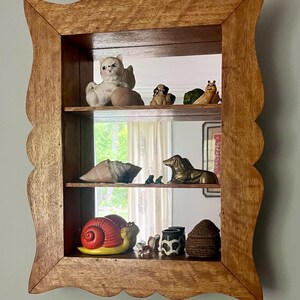May include: A wooden shadow box with a scalloped edge, displaying various ceramic figurines. The figurines include a cat, dogs, a snail, a seashell, and a brass dachshund. The box is mounted on a light blue wall.