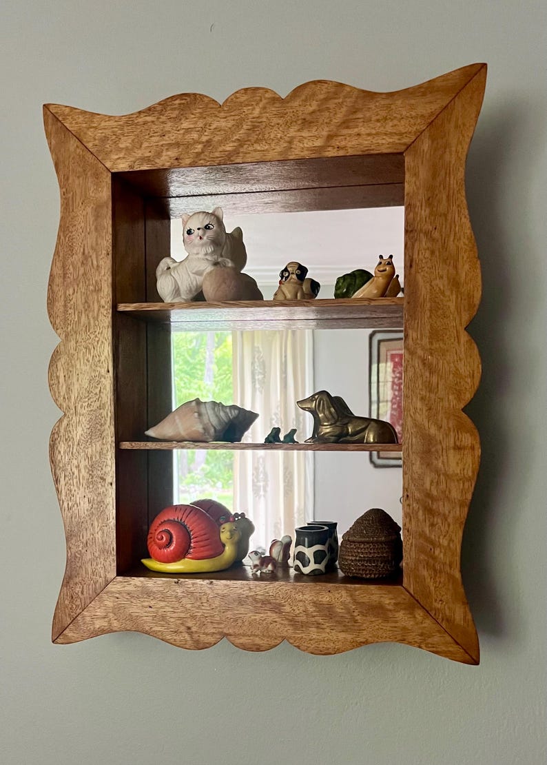 May include: A wooden shadow box with a scalloped edge, displaying various ceramic figurines. The figurines include a cat, dogs, a snail, a seashell, and a brass dachshund. The box is mounted on a light blue wall.