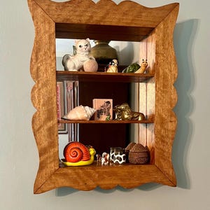 May include: A wooden, three-tiered shelf with a scalloped frame. The shelves display various figurines, including a white cat, a snail, a golden horse, and a small cow-print cup. The mirror backing reflects the items.