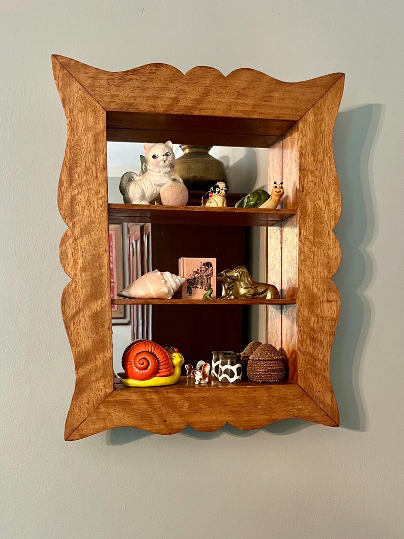 May include: A wooden, three-tiered shelf with a scalloped frame. The shelves display various figurines, including a white cat, a snail, a golden horse, and a small cow-print cup. The mirror backing reflects the items.
