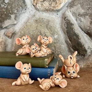 May include: A collection of six vintage ceramic mouse figurines in various poses. The mice are tan with large pink ears and black eyes. They are arranged on a wooden surface with a stone background and a stack of books.