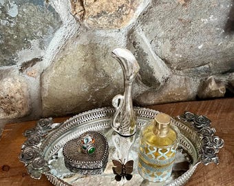 May include: A silver-toned oval vanity tray with rose-shaped handles, featuring a mirrored base. On the tray sits a silver swan-shaped bud vase, a heart-shaped trinket box with a green stone ring, a small metal butterfly, and a glass perfume bottle.
