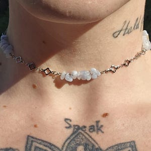May include: A delicate choker necklace featuring small, light blue gemstone chips strung on a silver-toned chain. The chain has a repeating pattern of open, four-petal flower shapes. The necklace is worn around the neck.