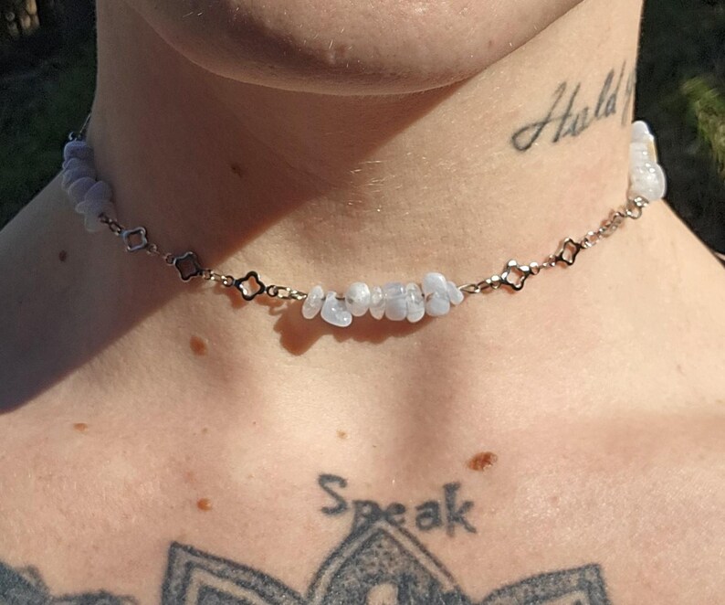 May include: A delicate choker necklace featuring small, light blue gemstone chips strung on a silver-toned chain. The chain has a repeating pattern of open, four-petal flower shapes. The necklace is worn around the neck.