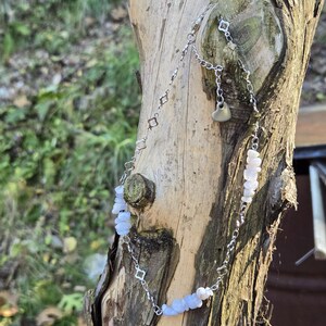 May include: A delicate silver necklace with a heart pendant and light blue gemstone beads. The necklace is draped over a weathered tree trunk, showcasing its intricate design and natural aesthetic. The necklace features a chain with small square-shaped links.